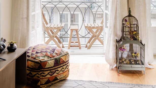 Ein Zimmer mit einem großen Fenster, einer Holzbänkl, einem farbenfrohen Pouf, einem Vogelkäfig und einem kleinen Tisch mit Dekorationen.