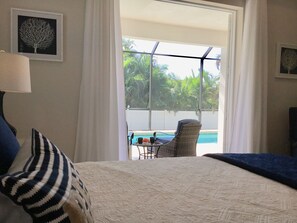 A bedroom with a bed, a chair, a table with a pool view, and a window with curtains.