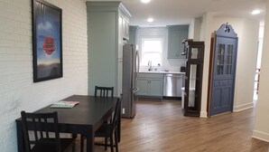 Une cuisine moderne avec des murs en briques blanches, un coin repas avec une table et des chaises en bois, et une porte bleue donnant sur une autre pièce.