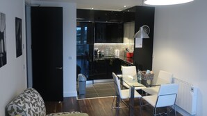 A modern living room with a glass dining table, white chairs, and a patterned sofa. A kitchen with black cabinets and a stainless steel range hood is visible through an open door. - Grand Canal Dock Corporate Apartment (Dublin)