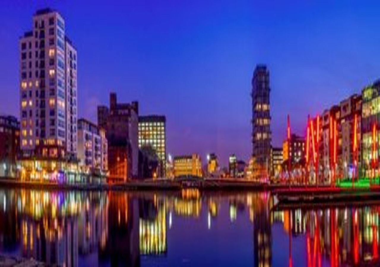 A nighttime cityscape with illuminated buildings reflected in a calm body of water.