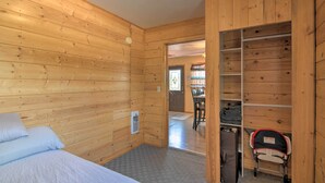 Una habitación con paneles de madera, una cama, un armario con estantes y un área de comedor visible a través de un umbral abierto.