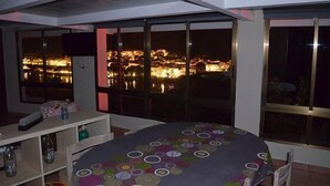 Een kamer met grote ramen met uitzicht op een stadsbeeld 's nachts, een ronde tafel met een kleurrijk tafelkleed, en een witte plank met diverse voorwerpen.