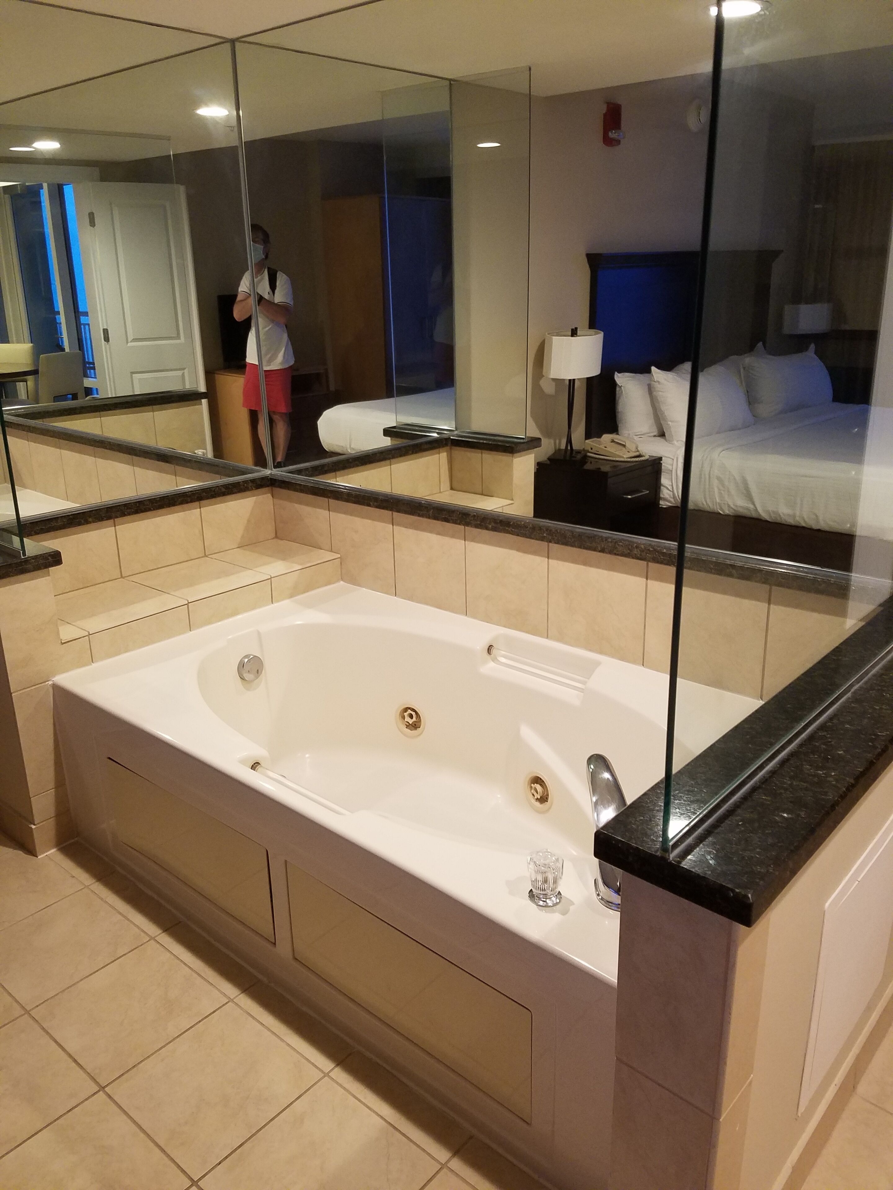 Une salle de bain moderne avec une grande baignoire, une cabine de douche en verre et un miroir qui reflète un lit et une table de chevet.