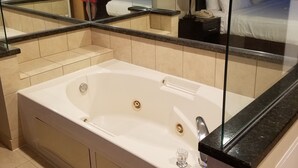 A modern bathroom with a large bathtub, a glass shower enclosure, and a mirror reflecting a bed and a nightstand.