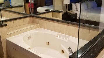 Une salle de bain moderne avec une grande baignoire, une cabine de douche en verre et un miroir qui reflète un lit et une table de chevet.