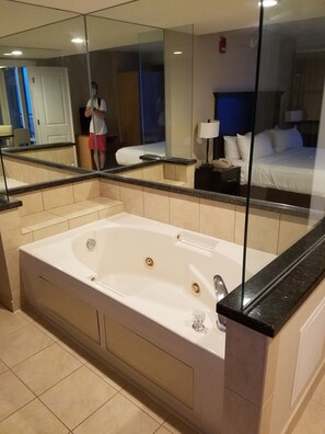 A modern bathroom with a large bathtub, a glass shower enclosure, and a mirror reflecting a bed and a nightstand. - Ocean Beach Club - Oceanfront 3BR Penthouse Suite. (Virginia Beach)