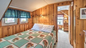 Une chambre avec un plafond lambrissé en bois, un lit recouvert d’une courtepointe à motifs floraux, deux fenêtres avec des rideaux bleus et un escalier menant à un étage supérieur.