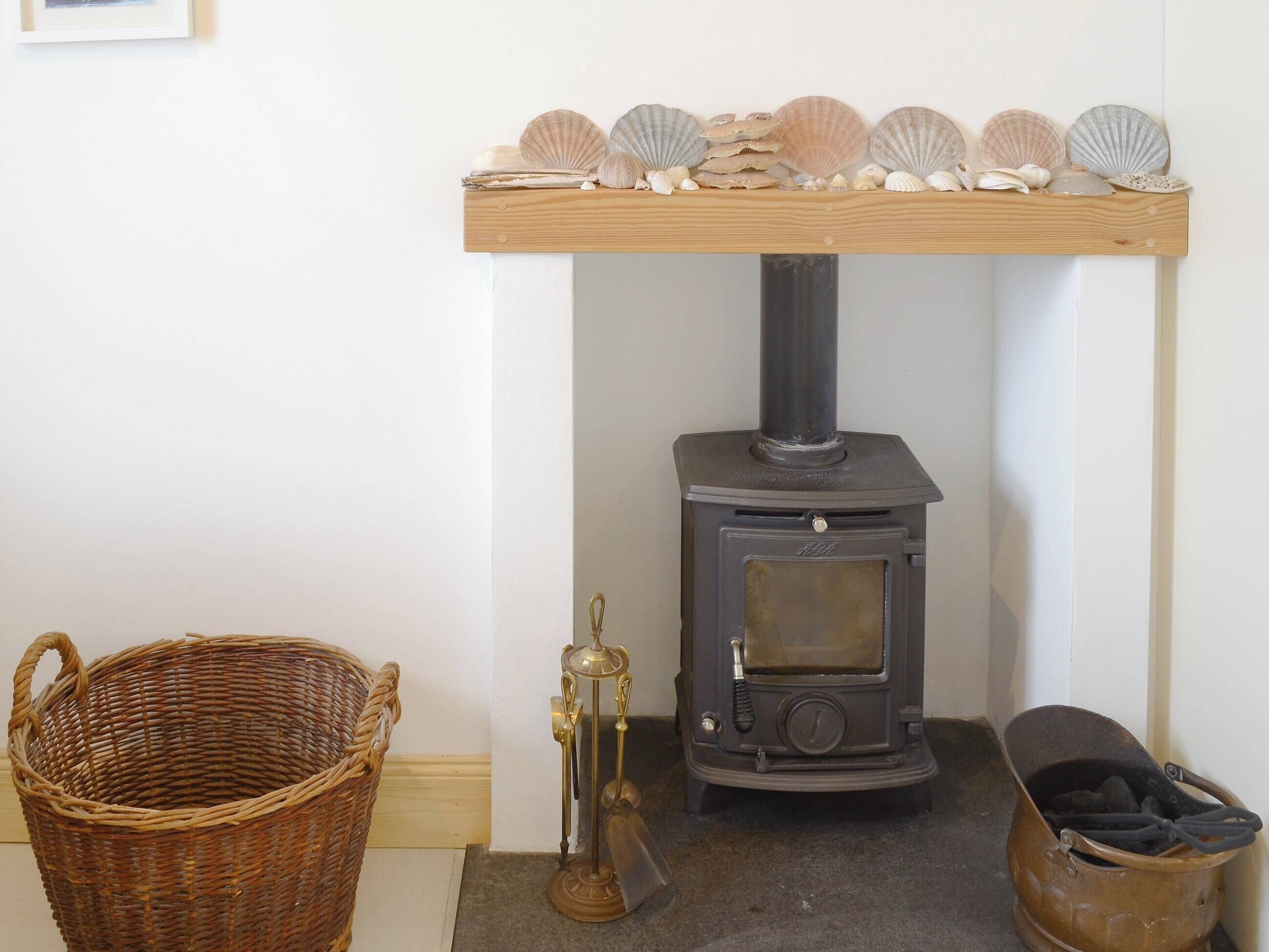 A wood-burning stove with a shelf above it holding seashells, a wicker basket, and a metal bucket.