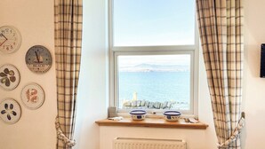 A room with a window overlooking the sea, a table with chairs, and wall clocks.