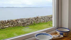 A view of a stone wall and grassy area through a window, with a bowl and plate on a table beside the window.