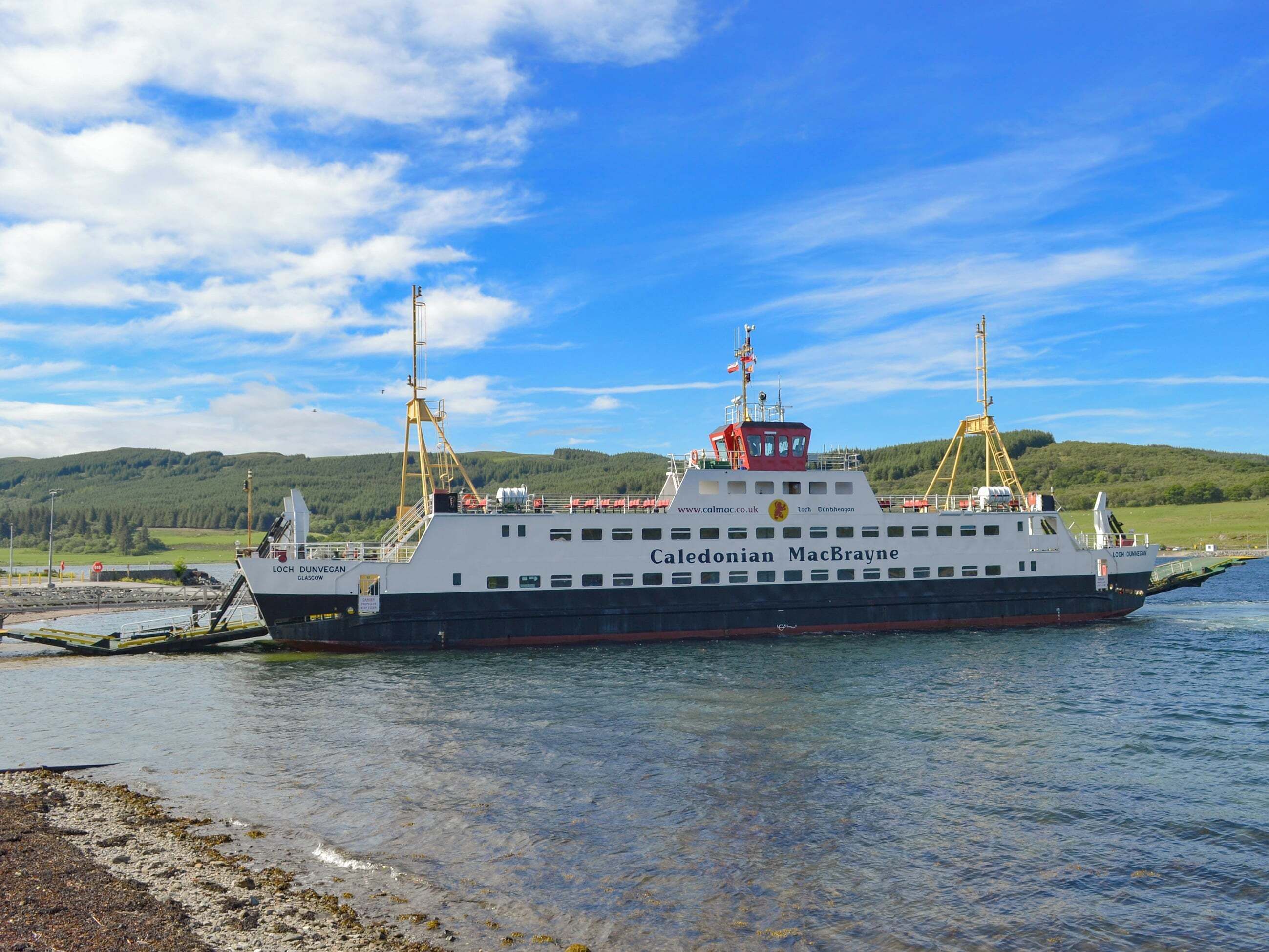 A large ferry named Caledonian MacBrayne docked at a pier with a clear sky and green hills in the background.