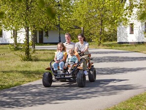 Eine Familie fährt mit einem Dreirad auf einem befestigten Weg. Im Hintergrund sind Bäume und Häuser zu sehen.