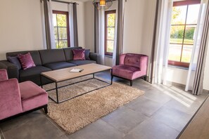 Una sala de estar con sofá, sillón, mesa de centro y ventanas grandes.