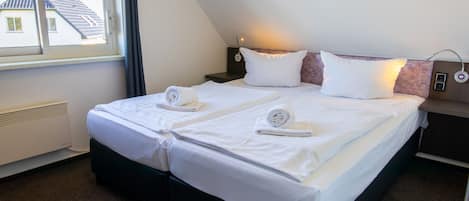 Una cama doble con ropa de cama y toallas blancas, una mesita de noche con una lámpara y una ventana con vistas a una casa.