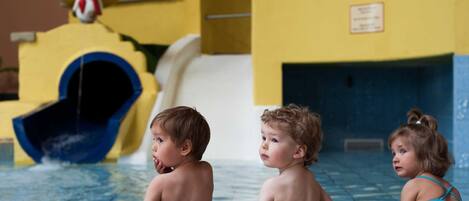 Children at a pool with a water slide and a building with arched windows in the background.