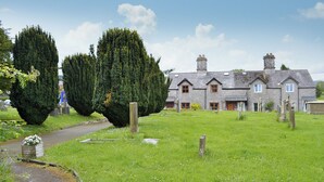 Ein Steinhäuschen mit einem Rasen und einem Weg zum Eingang, im Hintergrund ein Friedhof.