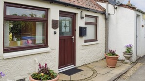 A white cottage with a brown door, two windows, and a satellite dish. There are two large flower pots on the porch. - Horseshoe Cottage is cosy and welcoming, perfect for a romantic break. (Freuchie, near Cupar)