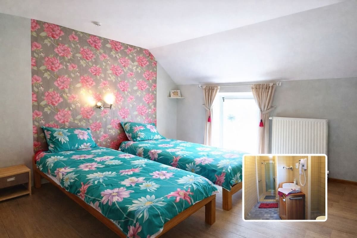 A bedroom with two beds, a window with curtains, a radiator, and a small bathroom with a shower.