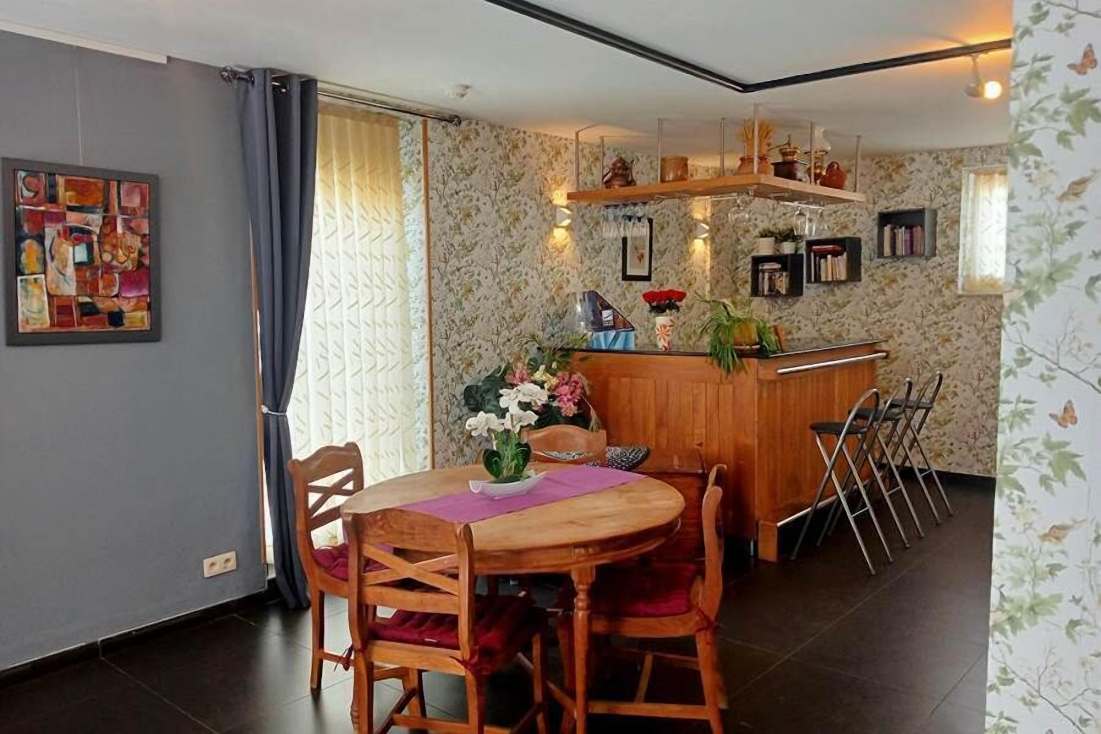 Uma área de refeições com mesa e cadeiras de madeira, um balcão de bar com banquetas e uma parede decorativa com quadros e plantas.