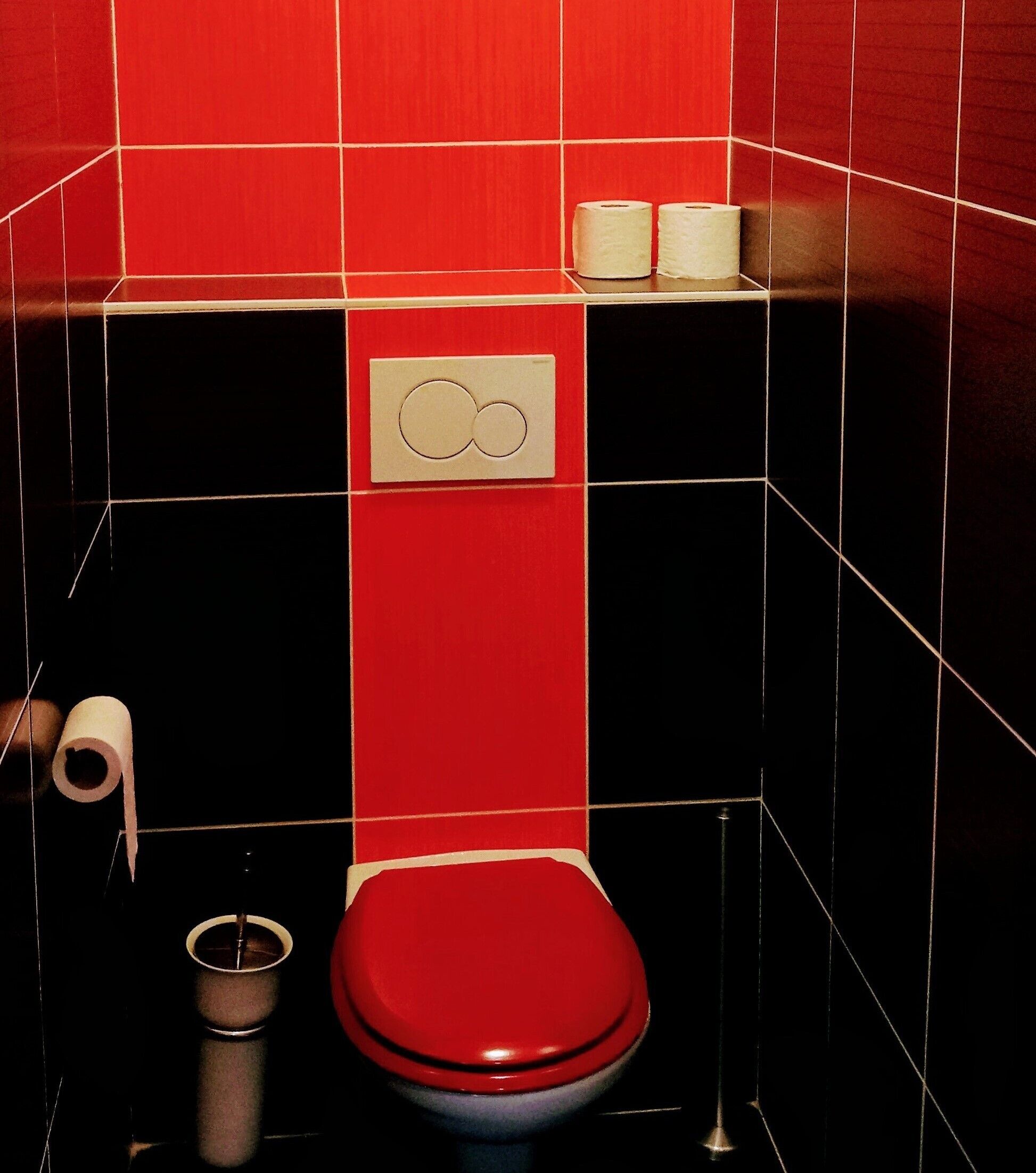Casa de banho com sanita vermelha, paredes revestidas a mosaico preto e vermelho, e duas embalagens de papel higiénico na parede.