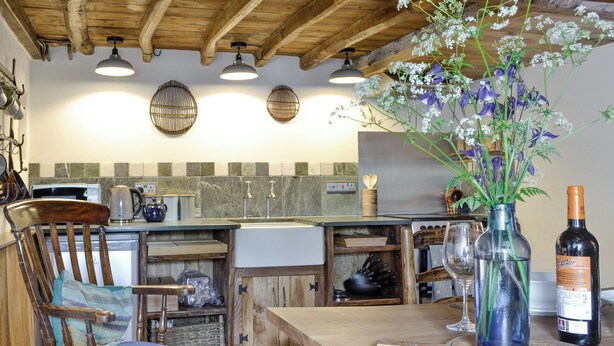 Una cocina rústica con vigas de madera, una mesa de comedor, una silla, un jarrón con flores y una botella de vino.