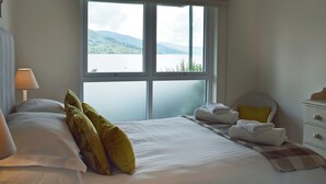 Un dormitorio con un ventanal grande, una cama con almohadas, una mesita de noche y una lámpara.