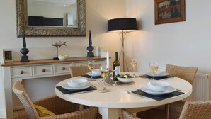 Un comedor con una mesa redonda preparada para cuatro personas, rodeada de sillas de mimbre. Un espejo y un cuadro enmarcado decoran la pared sobre la mesa.