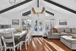 Amplia sala de estar con una mesa de comedor, sillas blancas, un sofá seccional gris y un ventanal con vista a árboles.