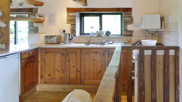 A kitchen with wooden cabinets, a stone accent wall, a window with natural light, and a built-in oven. - Woodland View-UK11826 (Sarnau, near Brecon)