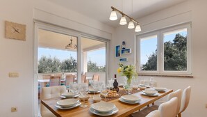 A dining area with a wooden table set for six, surrounded by white chairs. There are large windows providing natural light and a view of trees. - Cottage with barbecue on a plot of 2,500 sq.m. (Vinkuran)