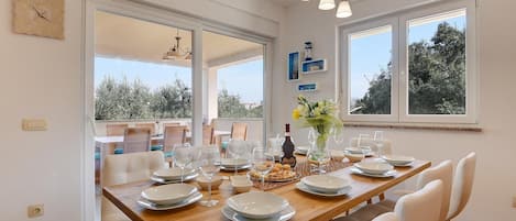 Uma sala de jantar com uma mesa de madeira posta para seis pessoas, rodeada por cadeiras brancas. Janelas amplas oferecem luz natural e vista para as árvores.