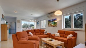A modern living room with orange sofas, a wooden coffee table, and a dining table. There is a kitchen area with a refrigerator and a dining table with chairs. Large windows provide natural light and views of trees. - Cottage with barbecue on a plot of 2,500 sq.m. (Vinkuran)