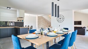 A modern dining area with a wooden table set for six, blue upholstered chairs, and a kitchen in the background.