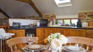 A dining area with a wooden table set for two, a kitchen in the background with stone walls, and a skylight above.