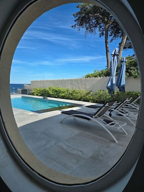 A circular view of a swimming pool with lounge chairs and umbrellas, a clear sky, and a distant view of the sea.