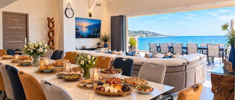 Comedor con vista al mar, un reloj en la pared y una variedad de comida y bebidas sobre la mesa.