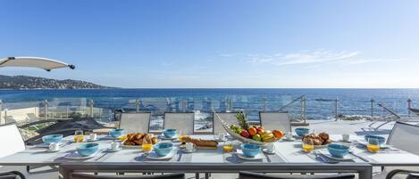 Una mesa preparada para el desayuno con vistas al mar, que incluye tazones, frutas y croissants.