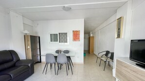 Ein modernes Wohnzimmer mit einem Glastisch, zwei grauen Stühlen, einem schwarzen Ledersofa, einem Holzschrank und einem Fernseher auf einem Standfuß.