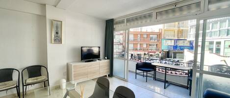 A modern living room with a flat-screen TV, a dining table with chairs, and a balcony with a view of the street and buildings.