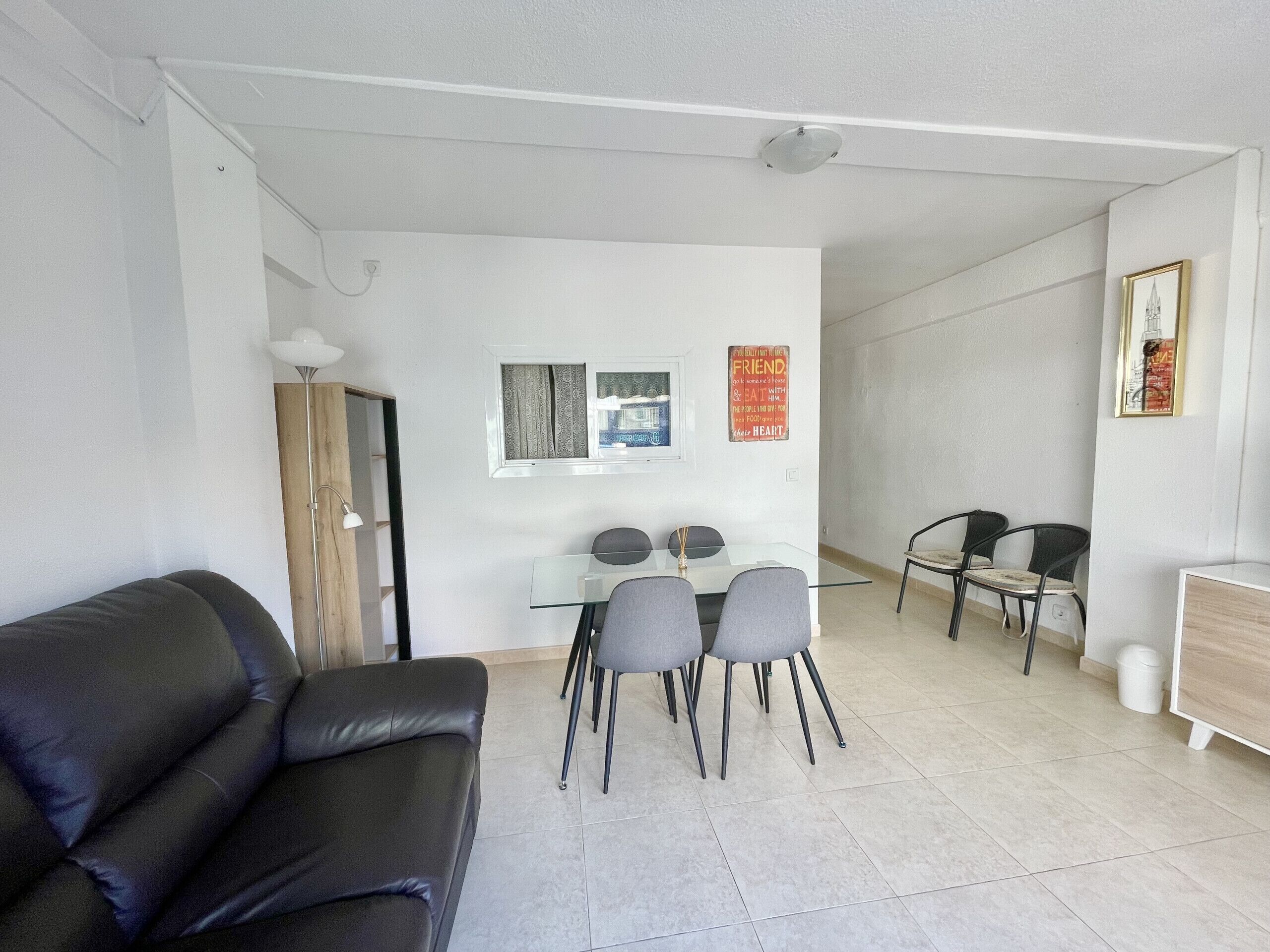 A modern living room with a black leather sofa, a glass dining table with four chairs, a small window with curtains, and a wall-mounted light fixture.