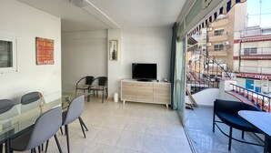 Ein modernes Wohnzimmer mit einem Glastisch, zwei Stühlen, einem Fernseher und einer Holzkommode. Durch die Glaswand ist ein Balkon mit Metallgeländer und Blick auf ein Gebäude zu sehen.