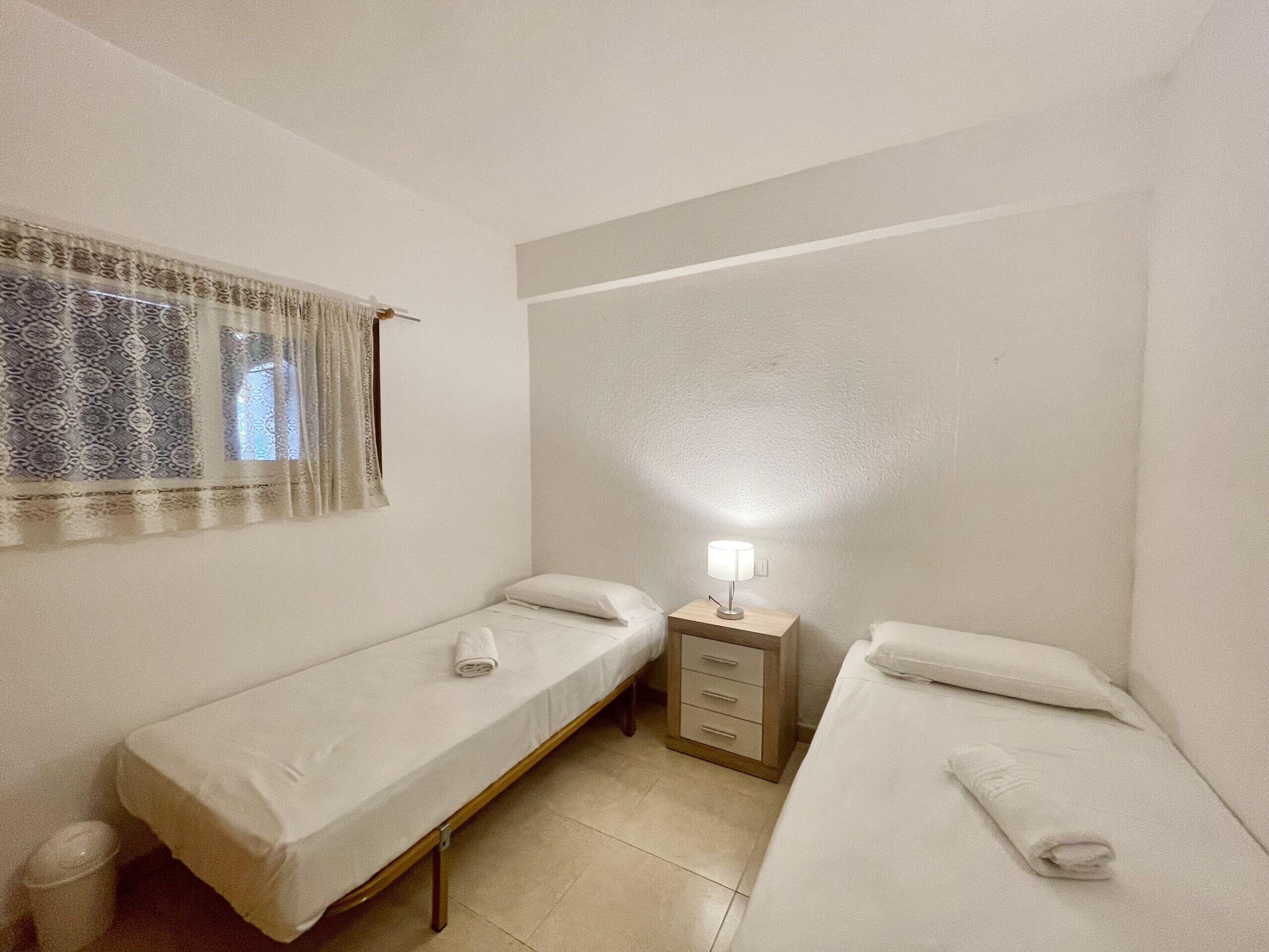 Une petite chambre propre, avec deux lits simples, une table de chevet avec une lampe et une fenêtre agrémentée de rideaux à motifs.