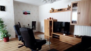 A modern living room with a black leather chair, a wooden entertainment unit with a TV, a glass coffee table, and a potted plant.