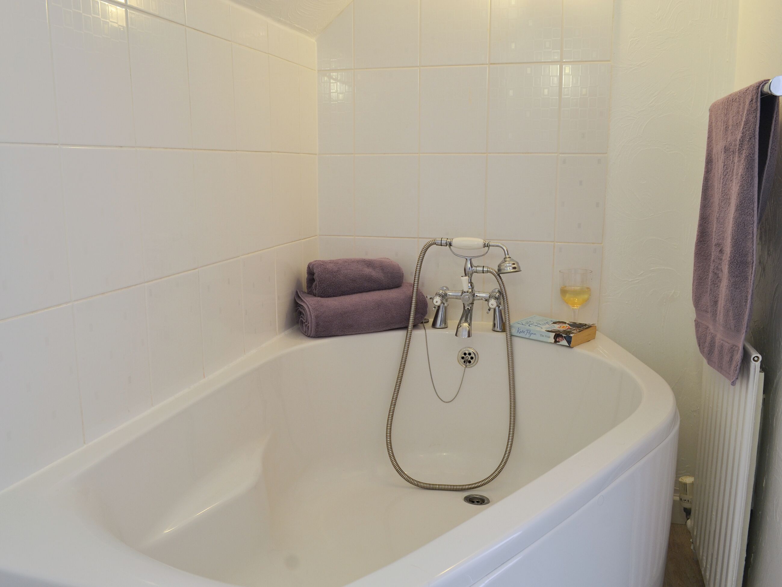 Une baignoire blanche, dotée d’un pommeau de douche à main et d’un robinet intégré. Une serviette violette est posée sur le bord, et un verre contenant un liquide jaune se trouve sur le bord de la baignoire.