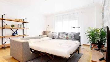 A modern bedroom with a sofa bed, a wooden shelf unit, a desk with a computer, and a large window with curtains.