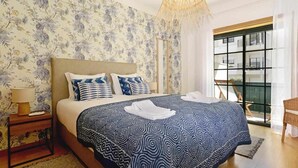 A bedroom with a patterned wallpaper, a large bed with a geometric bedspread, a bedside lamp, and a view of a building through a window.