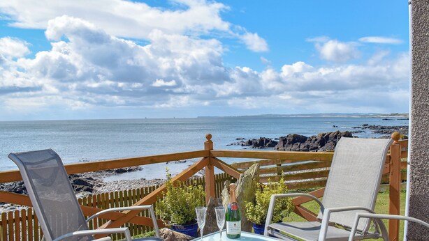 Una terraza de madera con asientos exteriores y una mesa, con vista a una costa rocosa y al mar.