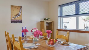 Una mesa de comedor dispuesta con platos, cubiertos y un cuenco de fruta. Hay sillas alrededor de la mesa, y una ventana con persianas a la derecha.