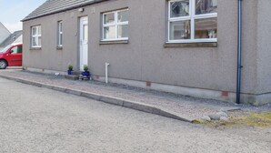 Un edificio de dos pisos con fachada gris, un coche rojo estacionado adelante y un pequeño jardín con macetas.